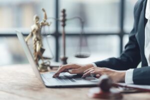 An attorney in a suit sitting at a desk typing on a laptop in front of scales of justice and lady justice.