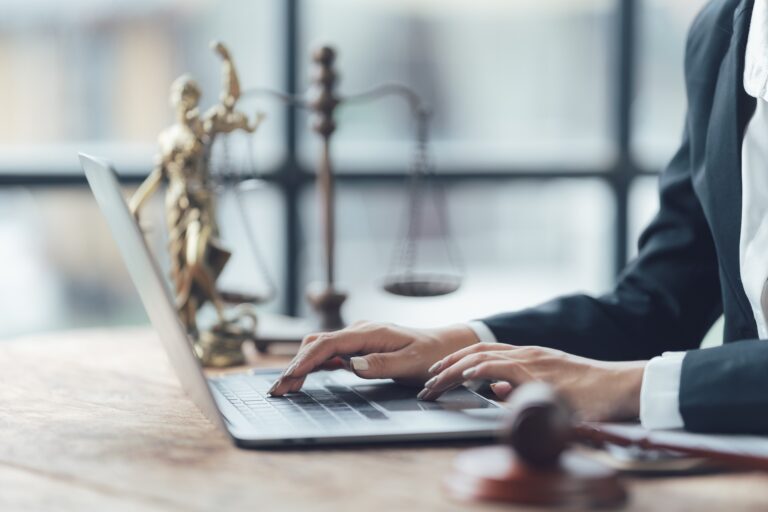 An attorney in a suit sitting at a desk typing on a laptop in front of scales of justice and lady justice.