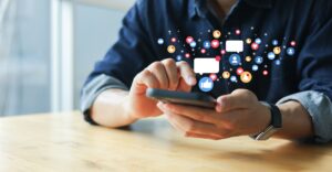 A person using a cell phone sitting at a table while social media graphics float above it.