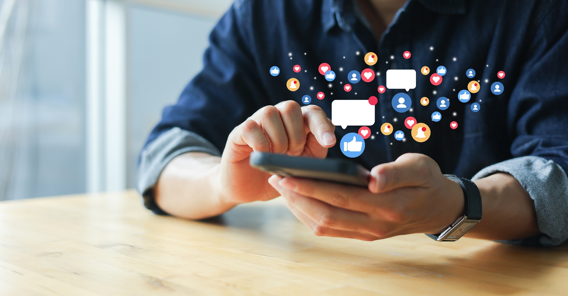 A person using a cell phone sitting at a table while social media graphics float above it.