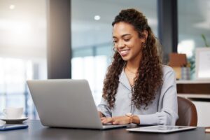 A woman sits behind a silver laptop at a desk smiling while looking at the screen and resting her hands on the keyboard.