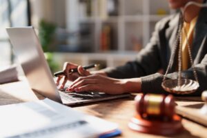 A person sits behind a laptop typing while holding a pen next to scales of justice.
