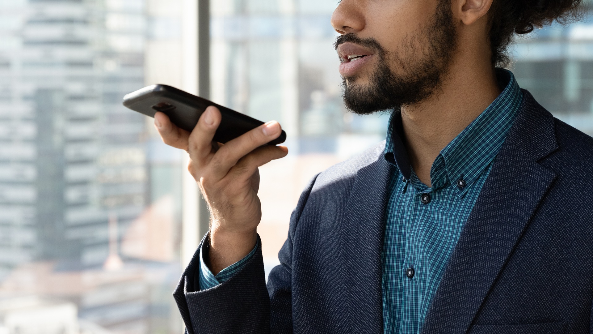 A man with a beard in a business suit holding a phone near his mouth in front of a city background.
