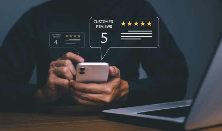 A woman holding a cell phone in front of her laptop looking at five star client reviews.