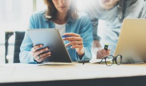 Two people behind a desk looking at a laptop while a pair of glasses and silver laptop sit next to them.
