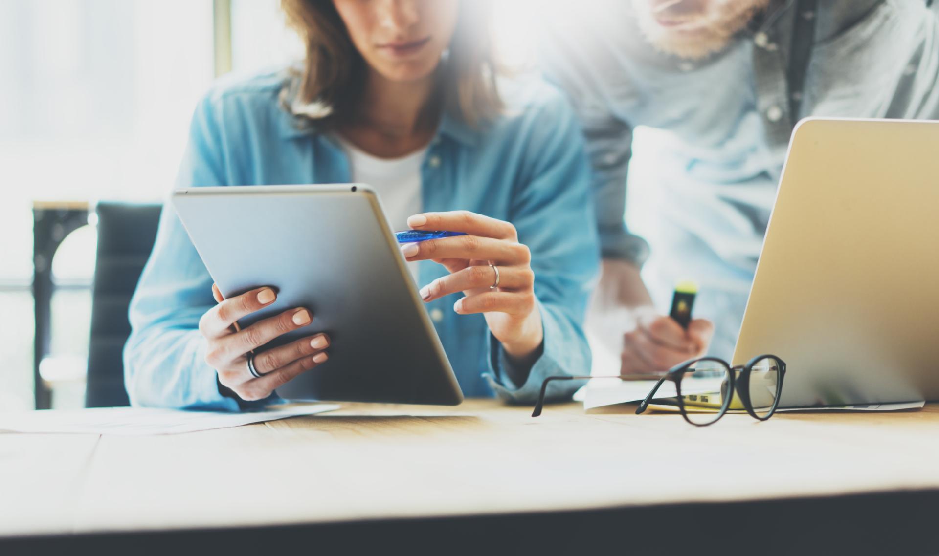 Two people behind a desk looking at a laptop while a pair of glasses and silver laptop sit next to them.