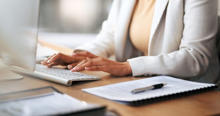A woman types on a keyboard at a desk next to a pad of paper with a pen resting on it.