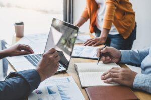 Three people sit and stand around a desk with laptops and pads of paper in a meeting.