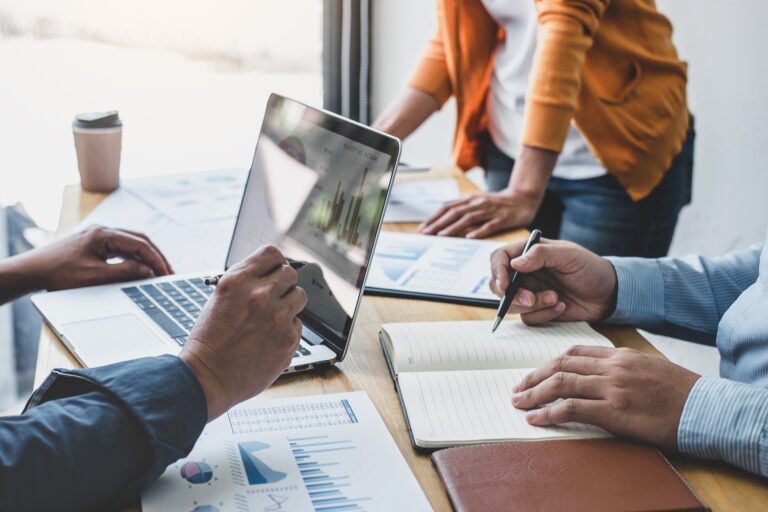 Three people sit and stand around a desk with laptops and pads of paper in a meeting.