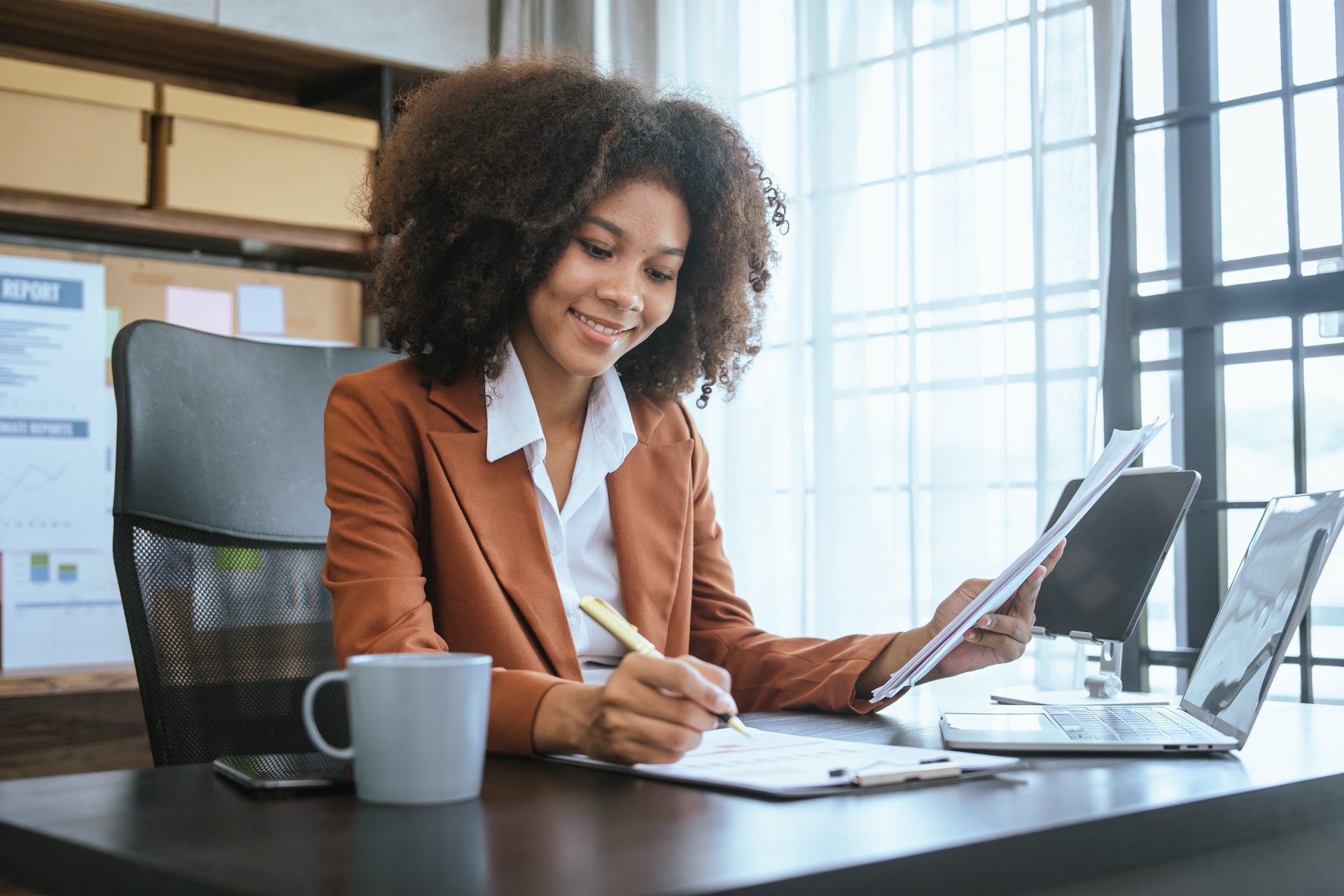 A business woman sitting at a desk writing on a paper and looking at a tablet in front of a laptop.