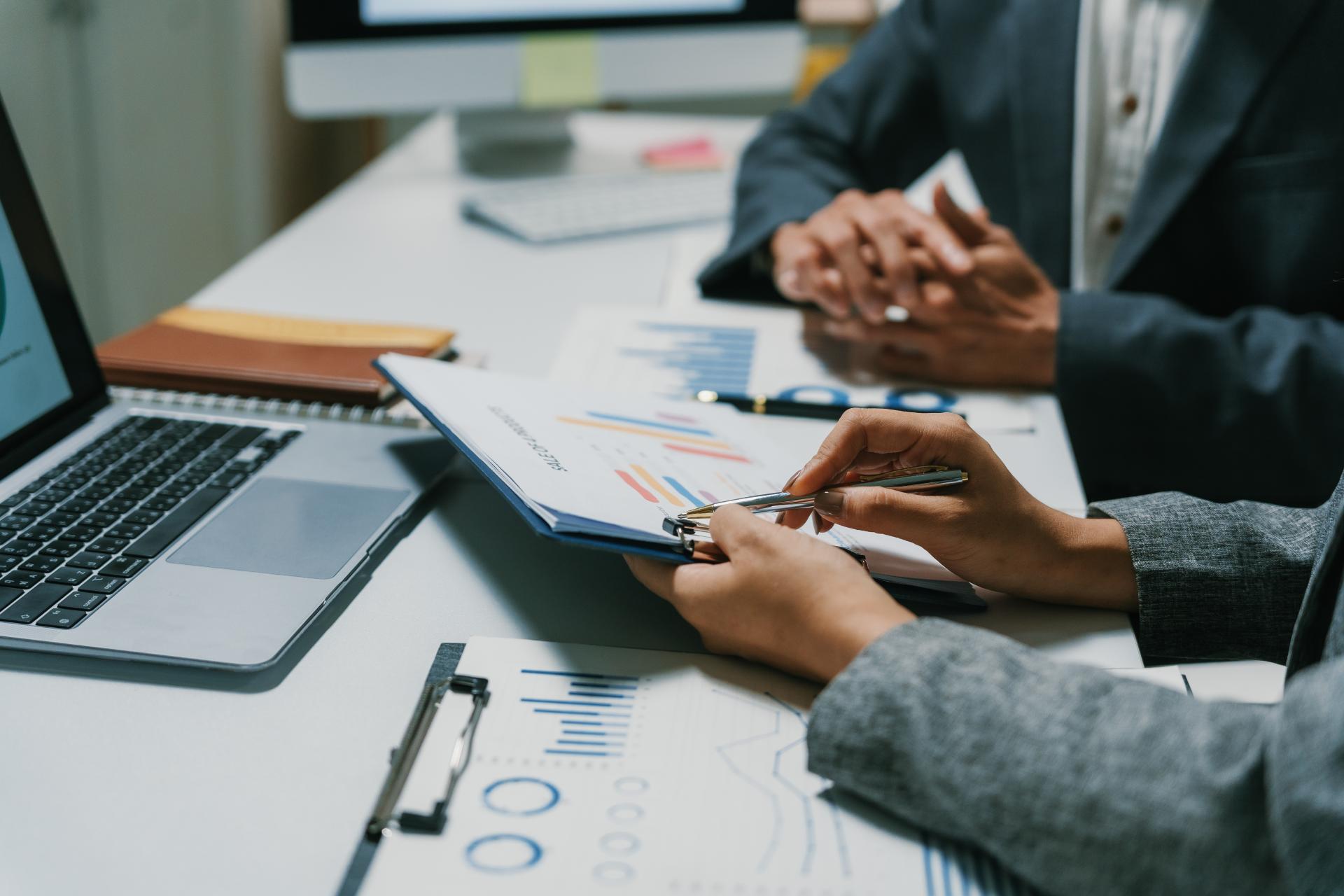 Two people sit at a desk looking at metrics on a clipboard in front of a silver laptop.
