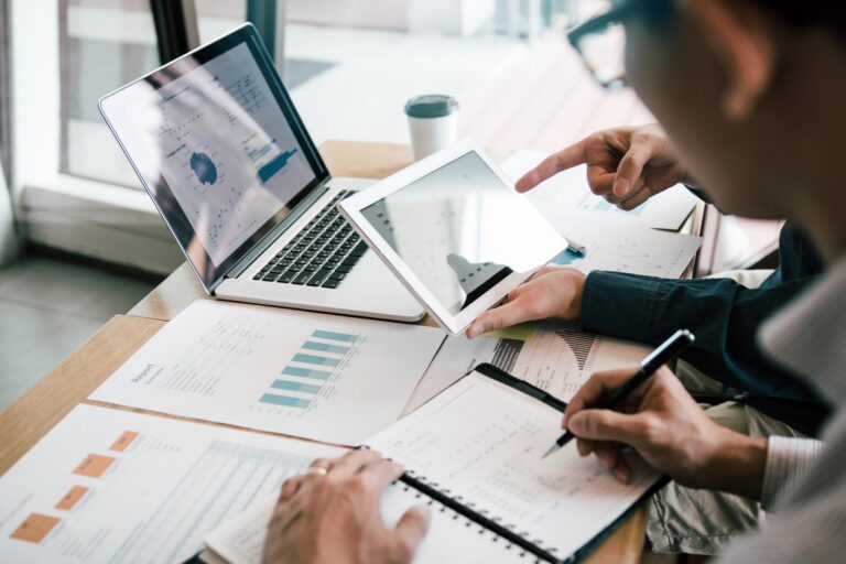 Two people sit at a desk analyzing data on papers, a tablet, and a laptop.
