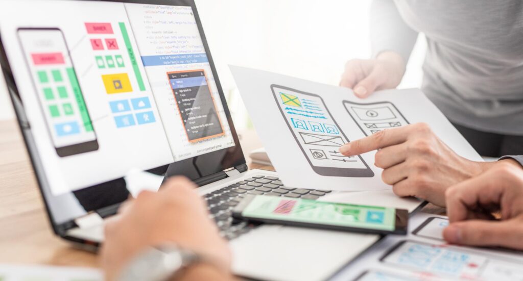 Two people sit in front of a laptop looking at designs on the screen while referencing images on paper.
